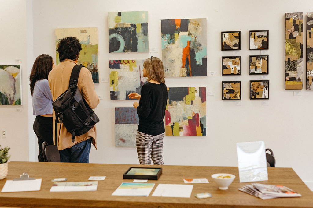 Abstract paintings in various sizes and colours line a white wall. Three people stand in front of them admiring the images