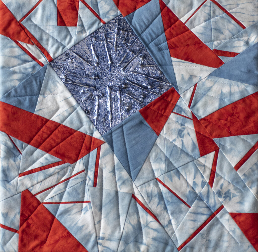 Abstract shapes in light blue and contrasting red. Off centre there is a darker blue square in a shimmering fabric.
