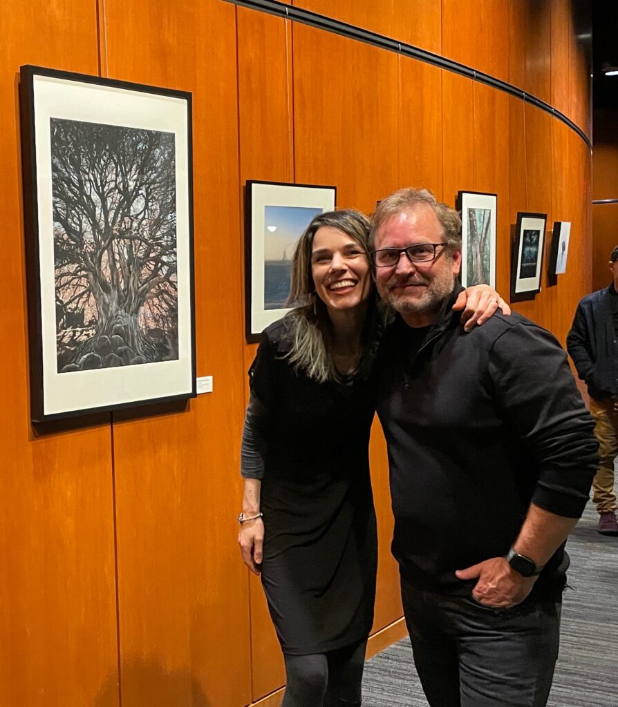 Victoria and Paul Newton at the opening of their exhibit 'Fusion Perspectives'. They are both smiling at the camera in front of their collaborative art pieces.