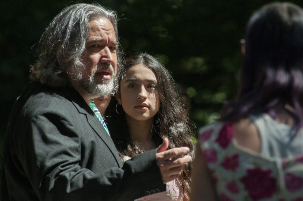 Performers from Shadows & Dreams Theatre Company perform a scene from Shakespeare's As You Like It. A man with shoulder length grey hair and a grey beard gestures towards the right of the image. A woman stands to the side of him, she has long dark hair and is wearing a pink dress.