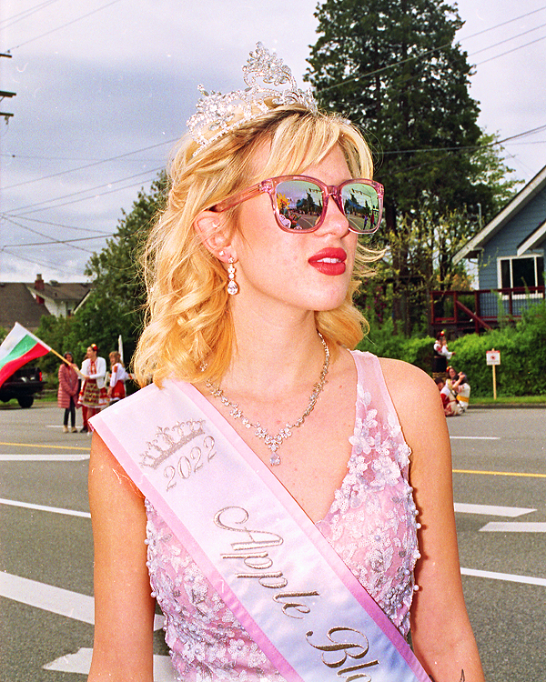 A young woman with blonde hair is standing in the street. She is wearing a pink dress, pink sunglasses and a pink sash. She also wears a crown and matching jewelry. She is not looking at the camera.