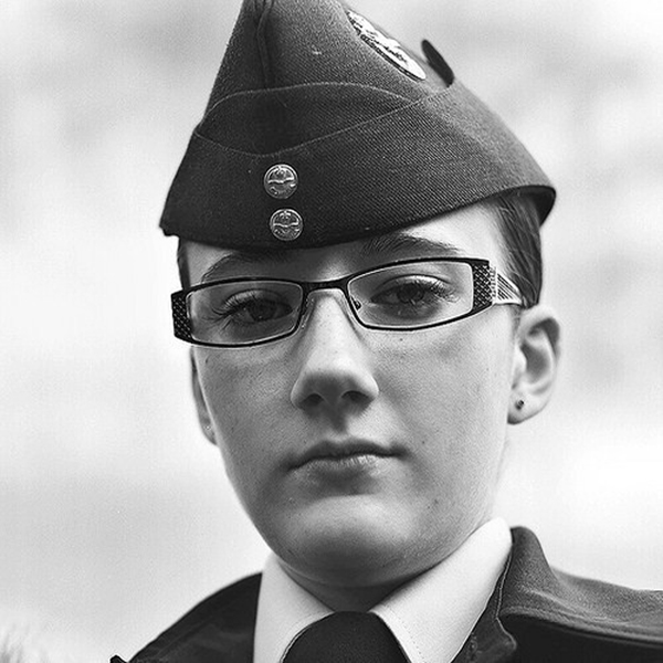 A black and white image of a girl wearing a uniform. Her hair is pulled up under a hat and she is wearing glasses. She does not smile at the camera.
