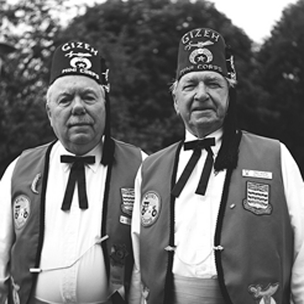 Two older men stand side by side. They are wearing the same uniform with hats, white shirts, black bowties, and vests. They both give small smiles to the camera.