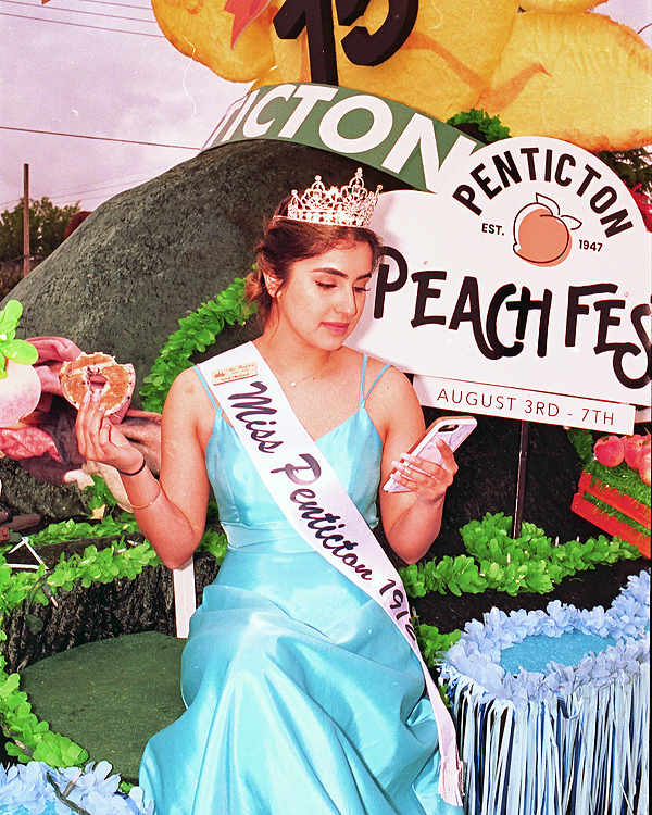 A girl in a bright blue dress sits on the edge of a green and yellow parade float. She is wearing a crown and a sash that reads "Miss Penticton". She is holding a half eaten bagel in one hand and a cell phone in the other.