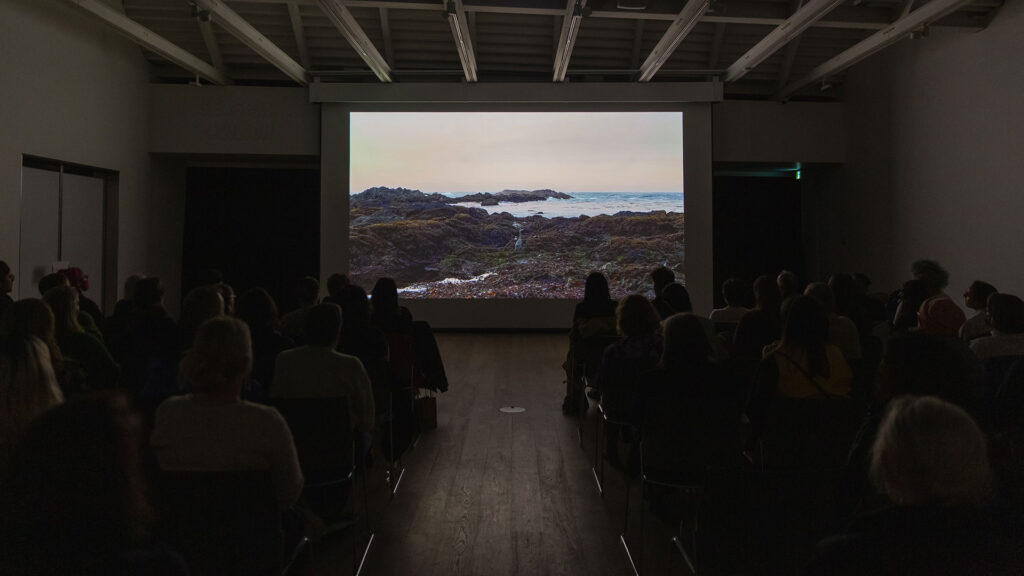 A full audience sits silhouetted against a projection screen featuring an image of a rocky ocean shore.