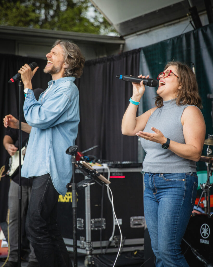 A man and woman both with brown hair and both wearing grey/blue shirts and jeans, are singing on stage