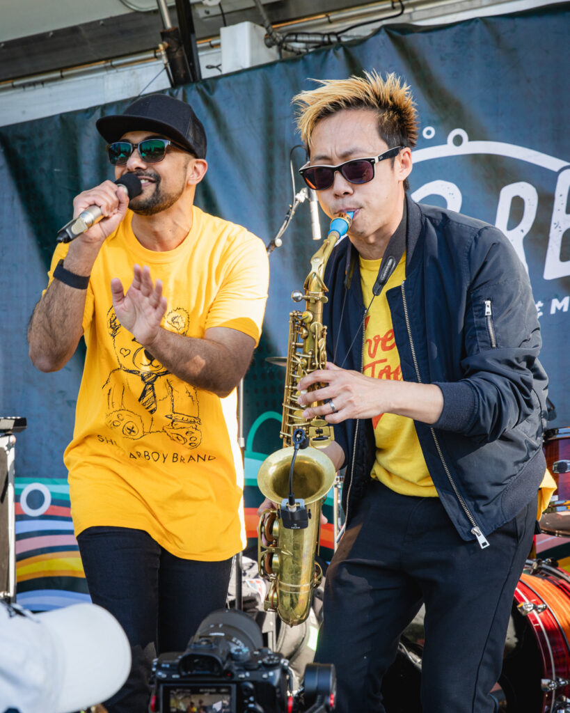 A man is singing into a mic, he is wearing a bright yellow shirt with a black hat, black pants and sunglasses. Next to him a man plays the saxophone, he has yellow tipped black hair and is wearing a yellow shirt beneath a black jacket and black jeans.
