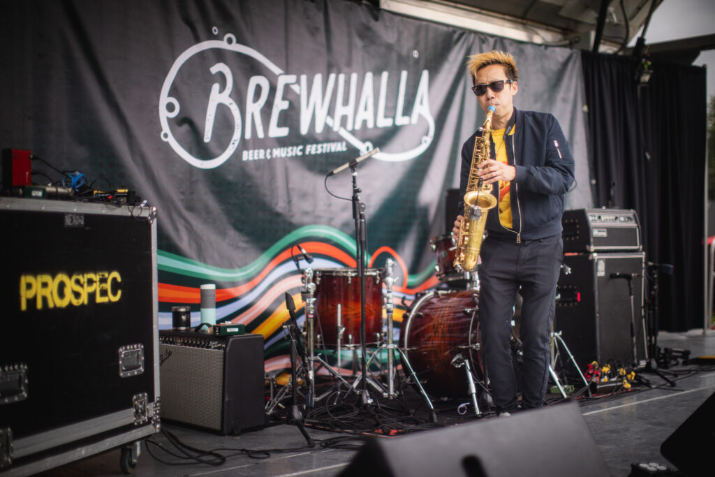 A man with blonde and black hair plays the saxophone on stage in front of the Brewhalla logo. He is wearing a black jacket and jeans with a yellow t-shirt.