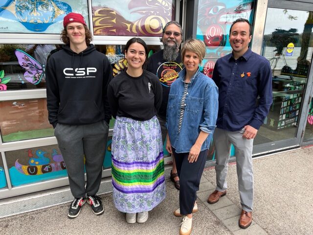 Executive Director Laura Grady stands with Indigenous Curator James Groening-Bluesky along with the 2023 Indigenous Artists in front of painted windows at River Market.