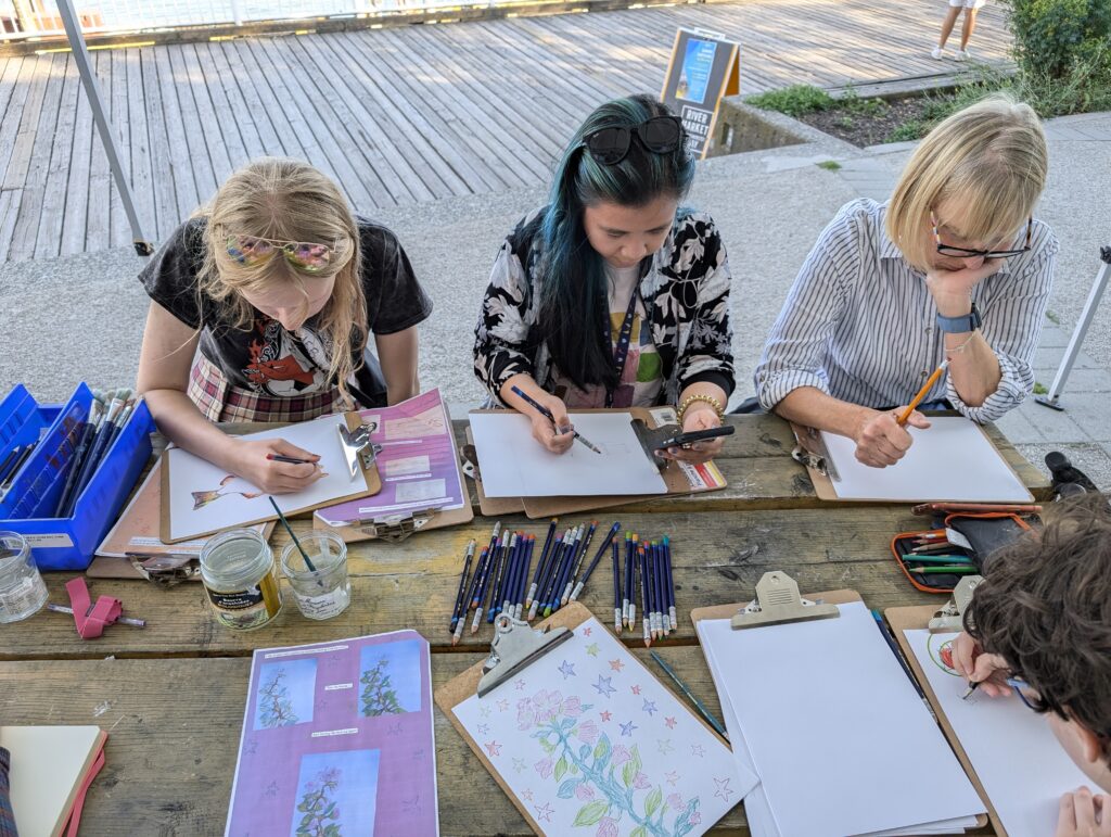 Participants sit around a wooden picnic table using pencils in sketchbooks.