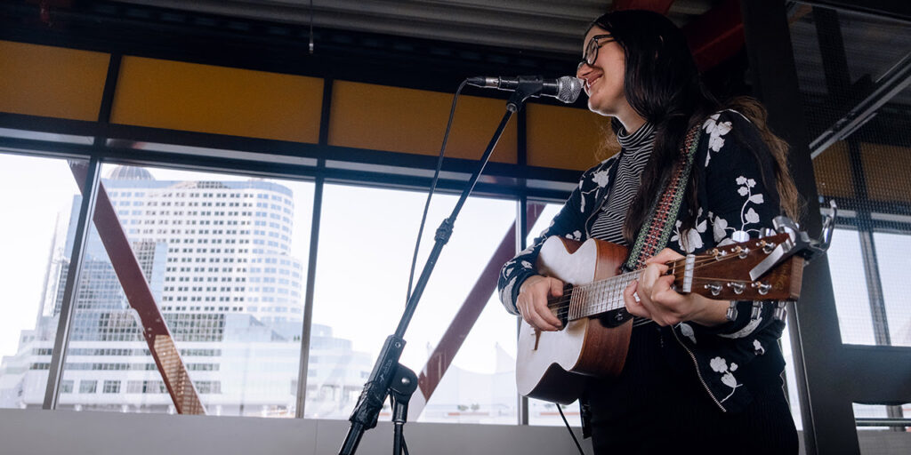 A woman is playing a guitar and singing into a mic in front of large windows looking out onto Canada Place in Downtown Vancouver.