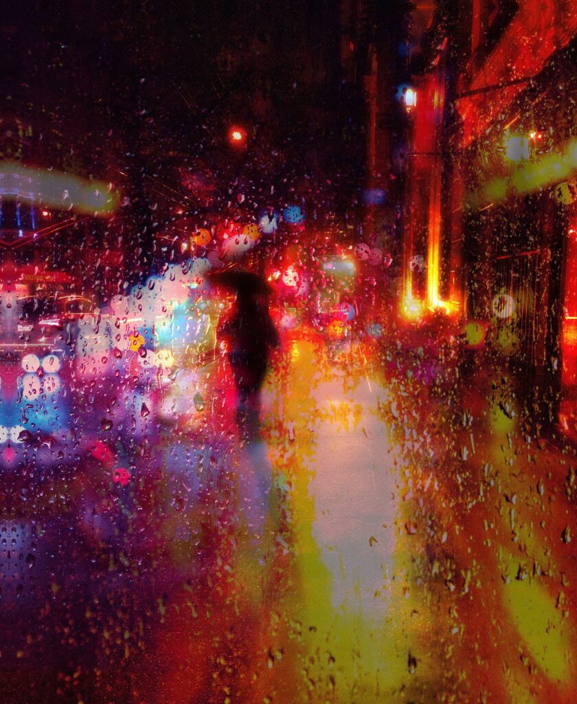 The shadowed silhouette of a person holding an umbrella walks into a street full of bright and vibrant lights that break into streaks through rain drops. 