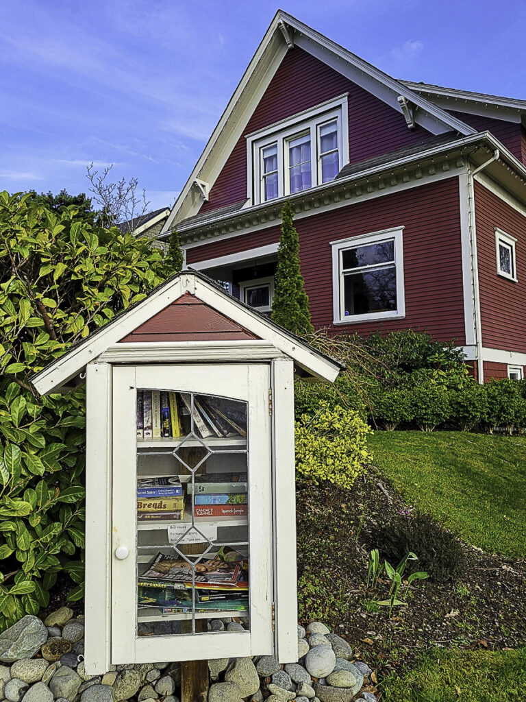 The free little library stands full of books in front of a red and white house.