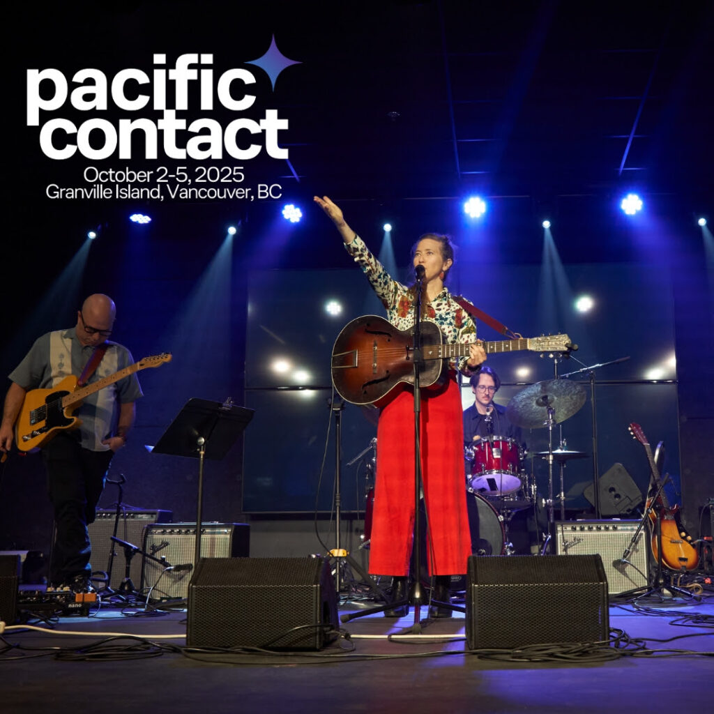 A woman wearing bright red pants and a cream patterned shirt gestures upwards as she sings into a mic while playing the guitar. A drummer and guitarist accompany her.