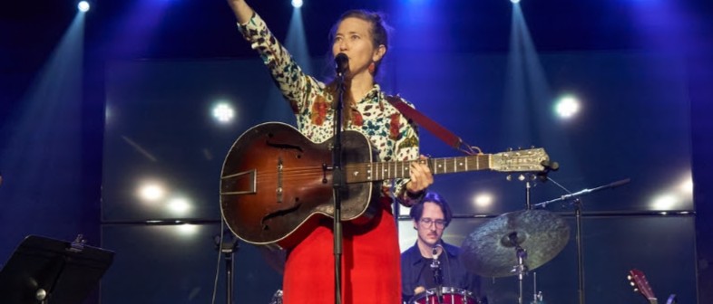 A woman in bright red pants and a cream pattered shirt gestures upwards as she sings into a mic.