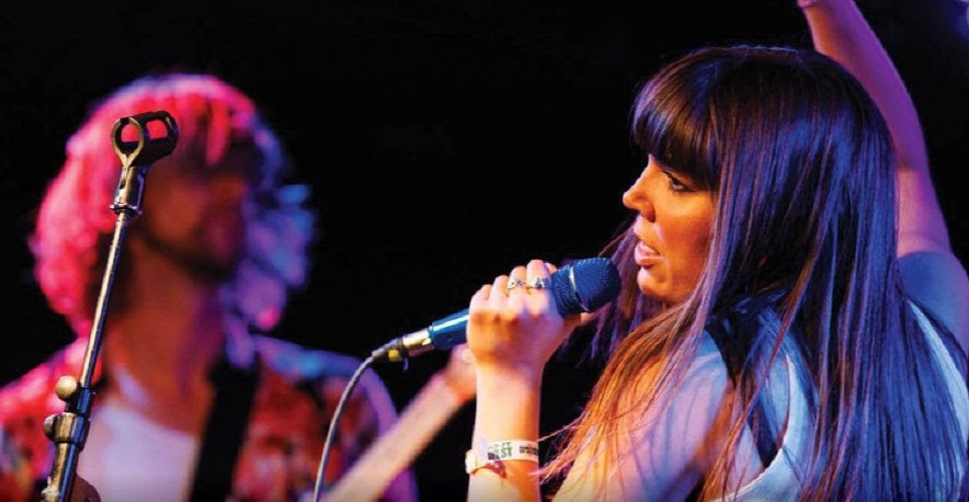 A woman with long brown hair sings into a microphone. She stands next to a man with a guitar.