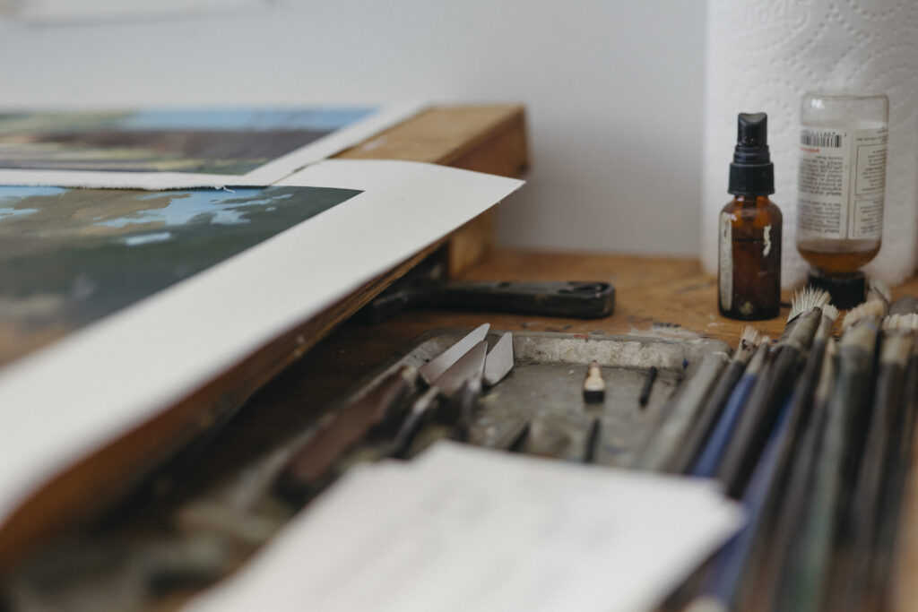 Two landscape paintings sit next to neatly aligned palette knives and brushes on a wooden tabletop