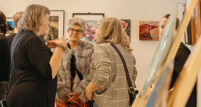 A trio stands in conversation next to a wall of easels on display in the Gallery at Queen's Park.