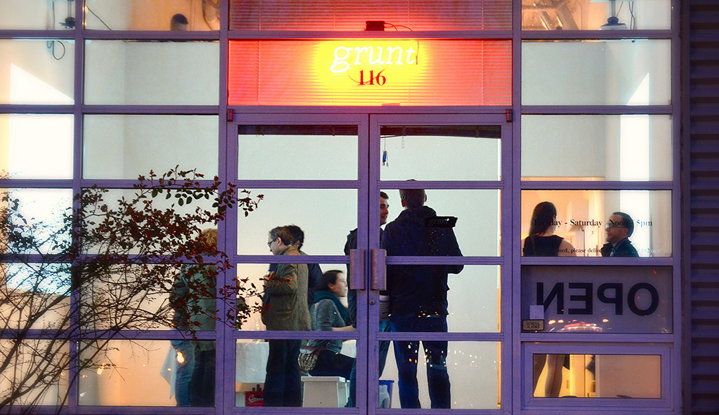 Through a glass door, people attend a party at the Grunt Gallery. The logo for the gallery is above the door in orange.