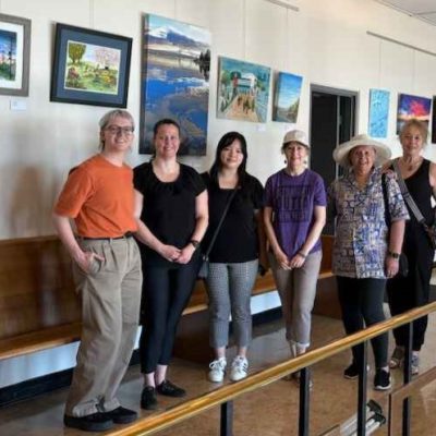 Members of Arts New West and New West Artists stand in front of a selection of brightly coloured paintings hung on the new Community Art Gallery wall at City Hall.