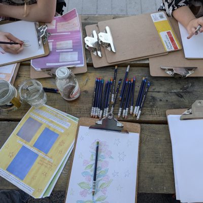 Sketchbooks and pencils are scattered across a wooden picnic table.