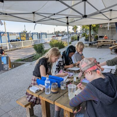 Participants sit around a wooden picnic table using pencils in sketchbooks.