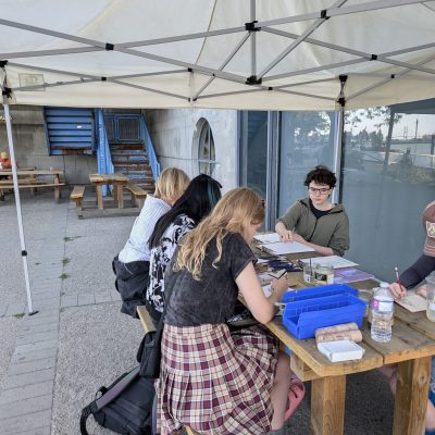 Participants sit around a wooden picnic table using pencils in sketchbooks.