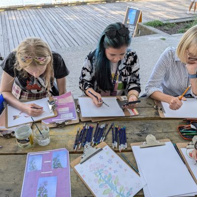 Participants sit around a wooden picnic table using pencils in sketchbooks.