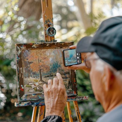 An older man holds up a cellphone to take a picture of a landscape painting.